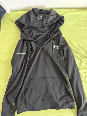 Svart träningshoodie med zip från Under Armour M - Säljer en svart hoodie från Under Armour med dragkedja vid halsen, stor huva och ficka framtill. Tillverkad i ett lätt och tekniskt material (HeatGear) som andas bra. Logga på bröstet och huvan. Perfekt för träning eller chill. Står M men passar bättre som S