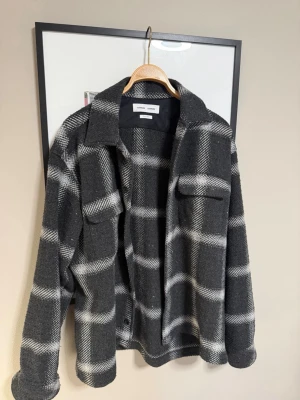 Grå rutig overshirt från Samsøe Samsøe - Snygg overshirt i grått och vitt rutmönster från Samsøe Samsøe. Tillverkad i italienskt tyg med klassisk krage, bröstfickor och avslappnad passform. Perfekt lager på lager-plagg för en trendig look.