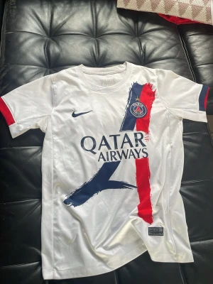 Vit PSG matchtröja Nike BARCOLA - Snygg vit PSG matchtröja från Nike med klubbmärke på bröstet och Qatar Airways-tryck. Tröjan har blå och röda penseldrag framtill, rund hals och korta ärmar med röda detaljer. Perfekt för fotbollsfans som vill sticka ut på läktaren eller planen.