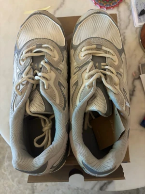 Vita och grå asics sneakers med meshdetaljer - Säljer ett para asics i vita och grå sneakers med meshdetaljer och klassisk snörning. Skorna har en sportig look med sula i vitt och grått samt ventilerande ovandel i mesh och syntet. Perfekta för dig som gillar stilrena och bekväma sneakers.