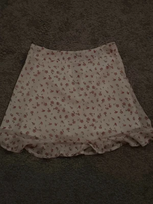 Blommig kortkjol med volangkant - Söt kortkjol i ljus färg med små rosa blommor och volangkant längst ner. Kjolen har en lätt och luftig känsla, perfekt för varmare dagar. Passar dig som gillar romantisk och feminin stil.