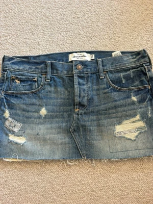 Blå jeanskjol från Abercrombie & Fitch - Snygg blå jeanskjol från Abercrombie & Fitch med slitna detaljer, rå kant nertill och patchade partier. Klassisk femficksmodell med bälteshällor och knappstängning. Perfekt för en avslappnad och trendig look.💓