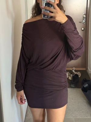 Mörkbrun offshoulder kortklänning - Säljer en mörkbrun kortklänning med offshoulder-design och långärmade ärmar. Klänningen har en loose passform upptill och smalnar av vid höften. Tillverkad i mjukt tyg, perfekt för en trendig och avslappnad look.