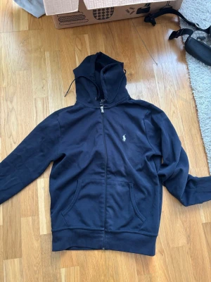 Marinblå zip hoodie från Polo Ralph Lauren - Snygg marinblå hoodie med dragkedja från Polo Ralph Lauren. Klassisk design med huva, fickor framtill och den ikoniska loggan broderad på bröstet. Perfekt för en avslappnad och stilren look.
