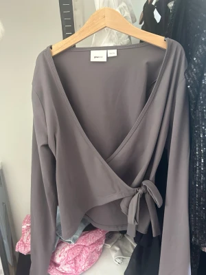 Grå omlottopp från Gina Tricot - Snygg grå omlottopp från Gina Tricot med långa ärmar och knyt i sidan. Toppen har en djup v-ringning och är tillverkad i ett mjukt, stretchigt material som sitter bekvämt på kroppen. Perfekt för att skapa en trendig och avslappnad look.