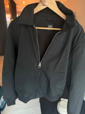 Svart bomberjacka från Zara - Snygg svart bomberjacka från Zara med dragkedja framtill och två fickor. Jackan har klassisk krage och ribbade muddar vid ärmslut och nederkant. Perfekt för dig som gillar en stilren och enkel look.