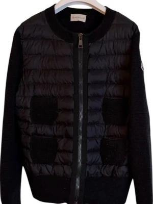 Svart cardiganjacka från Moncler - Svart cardiganjacka från Moncler med quiltad framsida i glansigt material och stickade ärmar samt detaljer. Dragkedja framtill och två stickade fickor. Moncler-logga på ärmen. Perfekt för lager-på-lager och snygg streetstil.