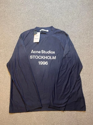 Blå långärmad topp Acne Studios - Mörkblå långärmad topp från Acne Studios med vit text 'Acne Studios STOCKHOLM 1996' på bröstet. Klassisk rund halsringning och avslappnad passform. Perfekt för en stilren och enkel look.