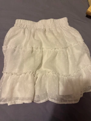 Vit volangkjol från FB Sister XS - Söt vit kortkjol från FB Sister i storlek XS. Kjolen har volanger och ett diskret prickigt mönster i tyget samt resår i midjan för skön passform. Perfekt för sommarens alla tillfällen och lätt att matcha med olika toppar.
