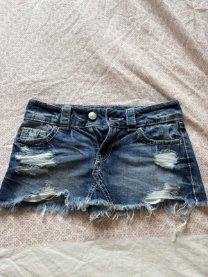 Blå jeanskjol med slitningar - Kort jeanskjol i blå denim med slitna detaljer och fransig nederkant. Kjolen har klassiska fickor fram och bak samt bälteshällor. Perfekt för en avslappnad och trendig look.