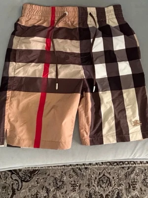 Rutig shorts med snörning - Snygga shorts med klassiskt rutigt mönster i beige, brunt, svart, vitt och en röd rand. Shortsen har elastisk midja med snörning och en bakficka. Perfekta för sommaren och ger en trendig look.