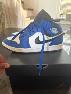 Air Jordan 1 Mid blå och vit - Snygga Air Jordan 1 Mid sneakers i blått, vitt och svart. Skorna har klassisk Nike Swoosh på sidan, Air Jordan-logga på ankeln och svart yttersula. Ovandelen är i läder med perforerad tåbox och blå skosnören. Perfekta för dig som gillar streetwear och ikonisk design.