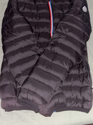 Svart dunjacka från Moncler - Säljer en svart dunjacka från Moncler med quiltad design och klassisk logotyp på ärmen. Jackan har dragkedja med rödblå detalj framtill och elastiska muddar vid ärmslut och nederkant. Perfekt för kalla vinterdagar.
