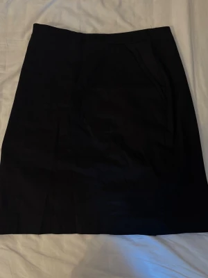 Svart enkel kjol med ficka - Stilren svart kjol i enkel design med praktisk ficka framtill. Kjolen har en rak passform och är perfekt för dig som gillar minimalistisk stil. Materialet känns mjukt och lätt, vilket gör den bekväm att bära hela dagen.