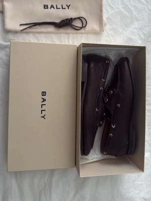 Bruna seglarskor i läder från Bally - Snygga seglarskor från Bally i mörkbrunt läder med klassisk snörning och guldfärgade öljetter. Skorna har en stilren design och låg sula, perfekta för en avslappnad men ändå uppklädd look. Kommer i originalkartong och med dustbag. Aldrig använda