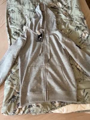 Grå zip hoodie från Stone Island, pris kan diskuteras. - Snygg grå hoodie från Stone Island med dragkedja framtill och två stora fickor. Klassisk huva med dragsko och ribbade muddar vid ärmar och nederkant. Tillverkad i mjukt material som känns skönt mot huden. Perfekt för en avslappnad stil.
