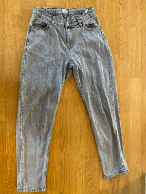Grå jeans med hög midja - Säljer ett par grå mom jeans med hög midja och raka ben. Jeansen har klassiska fem fickor, bälteshällor och knappgylf. Tillverkade i denim med en avslappnad passform som ger en cool vintagekänsla.
