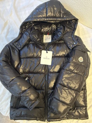 Svart dunjacka med huva från Moncler - Snygg svart dunjacka från Moncler med glansig finish och huva. Jackan har dragkedja framtill, fickor på sidorna och Moncler-logga på ärmen. Perfekt för kalla vinterdagar och riktigt trendig look. Fick den i present men den passade inte min stil så säljer den, har aldrig använt den! 