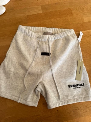 Grå shorts Essentials Fear of God - Snygga ljusgrå shorts från Essentials Fear of God med svart logga på benet. De har snörning i midjan och är tillverkade i mjuk bomull, perfekta för sommaren. Klassisk och avslappnad passform.