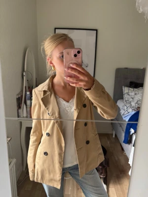 Beige trenchcoat med knytskärp - Stilren beige trenchcoat med dubbelknäppning och klassisk krage. Jackan har ett matchande knytskärp i midjan och svarta knappar. Passar perfekt till jeans eller kjol för en trendig look. 