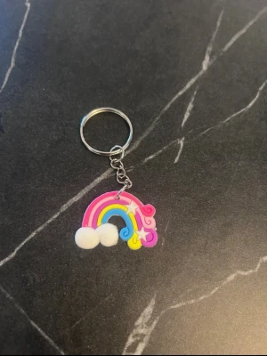 Nyckelring med regnbåge och moln - Färgglad nyckelring med en regnbåge i rosa, blått och gult, dekorerad med vita moln och en liten stjärna. Tillverkad i plast och metall. Perfekt att hänga på väskan eller nyckelknippan för en lekfull touch.
