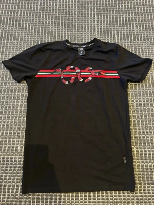 Svart t-shirt med ormtryck från Black Squad - Svart t-shirt från Black Squad med slim fit-passform. Framtill finns ett tryck med en röd och grön rand samt en orm i mitten. Perfekt för dig som vill sticka ut med en cool detalj. Klassisk rund hals och korta ärmar.