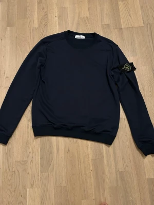 Mörkblå sweatshirt från Stone Island - Snygg mörkblå sweatshirt från Stone Island med klassisk rund halsringning och ribbade muddar. Tröjan har den ikoniska Stone Island-loggan på vänster ärm och är tillverkad i mjukt material för en clean och stilren look. Använd 2 gånger och pris kan diskuteras 