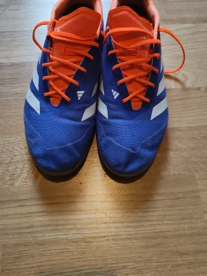 Adidas predator elite inomhus skor  - Snygga fotbollsskor i blått med vita detaljer och orange snörning. Skorna har en låg profil och ovandel i mesh-liknande material för bra ventilation. Yttersulan är vit och mönstrad för bra grepp på planen. Perfekta för dig som vill sticka ut på fotbollsplanen.