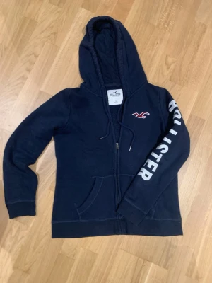 Mörkblå Hollister hoodie med dragkedja - Mörkblå hoodie från Hollister med dragkedja framtill och stor huva. Stora vita bokstäver med 'HOLLISTER' på ena ärmen och klassisk logga på bröstet. Mjuk bomullsblandning och kängurufickor gör den både snygg och bekväm. 