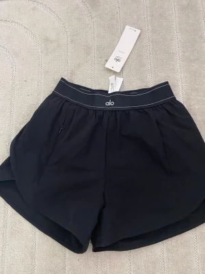 Svarta träningsshorts från Alo M - Snygga svarta träningsshorts från Alo med elastisk midja och vit kontrastsöm. Shortsen har en sportig look, rundade benslut och diskret logga framtill. Perfekta för gymmet eller löprundan.