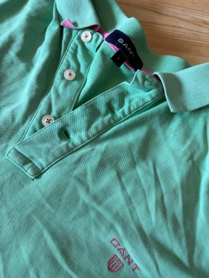 Grön pikétröja från Gant - Snygg grön pikétröja från Gant med klassisk krage och tre knappar framtill. Tröjan har korta ärmar och Gant-logga broderad i rosa på bröstet. Perfekt till sommaren och enkel att matcha med jeans eller shorts.