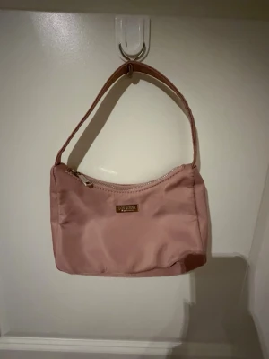 Rosa axelremsväska från Don Donna - Snygg rosa axelremsväska från Don Donna i ett glansigt tygmaterial. Väskan har en enkel, rektangulär form med dragkedja upptill och ett metallmärke framtill. Perfekt för dig som gillar stilrena accessoarer med en feminin touch.
