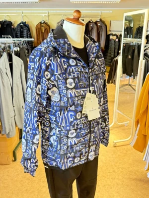Blå mönstrad vindjacka Moncler - Snygg vindjacka från Moncler i blått och vitt med grafiskt mönster och logotyper över hela jackan. Jackan har huva, dragkedja framtill och elastiska muddar vid ärmarna. Perfekt för dig som vill sticka ut med en unik streetwear-look.