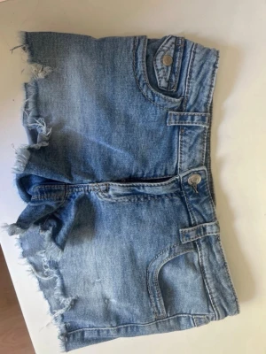 Ljusblå jeansshorts med fransar - Säljer ett par ljusblå jeansshorts med råa, fransiga benslut och klassiska fickor både fram och bak. Shortsen har bälteshällor och knappstängning. Perfekta för varma dagar och en avslappnad stil.