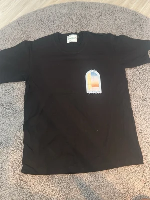 Svart t-shirt med tryck Casa Blanca - Snygg svart t-shirt med färgglatt tryck på bröstet där det står Casa Blanca. T-shirten har klassisk passform och rund hals. Perfekt till jeans eller shorts för en chill look.