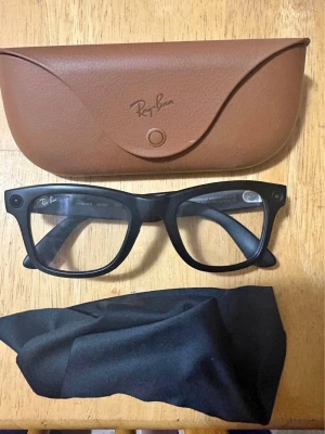 Svarta Ray-Ban solglasögon Wayfarer - Ray-Ban Meta smartglasögon i nyskick, köpta för två månader sedan och använda i mindre än två timmar. Glasögonen har mattsvarta bågar, Transitions-glas och inbyggda kameror. AI-funktioner finns integrerade för handsfree-användning. Originalets bruna laddningsfodral, svart rengöringsduk samt den öppnade originalkartongen medföljer. 