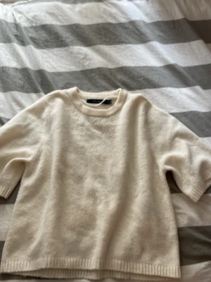 Krämvit stickad tröja - Mysig krämvit stickad tröja med rund hals och halvlånga ärmar. Tröjan har en rak passform och ribbstickade muddar vid ärmslut och nederkant. Perfekt att styla lager på lager under kyliga dagar.