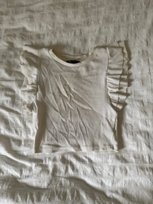 Vit ribbad topp med volangärm från Zara - Supersöt vit ribbad topp från Zara med volangdetaljer på ärmarna. Toppen har rund halsringning och är tillverkad i mjukt bomullsmaterial. Perfekt för dig som gillar en feminin och trendig stil.
