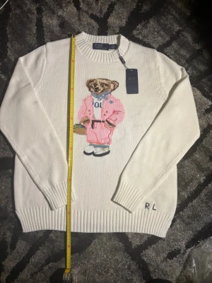 Vit stickad tröja med björnmotiv RL - Vit stickad tröja från Polo Ralph Lauren med ikoniskt björnmotiv på bröstet, där björnen bär rosa outfit och RL-broderi vid ärmslutet. Tröjan har rund hals, ribbade muddar och är tillverkad i mjukt material. Perfekt för dig som gillar statement-plagg.