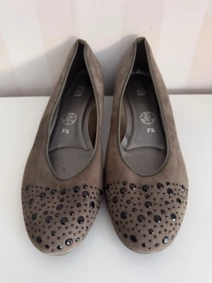 Beige ballerinaskor med nitar Ara st.37,5 - Snygga beige ballerinaskor med rund tå och dekorativa svarta nitar framtill. Skorna har en klassisk form och är tillverkade i mockaliknande material. Perfekta för dig som gillar en stilren look med en twist.