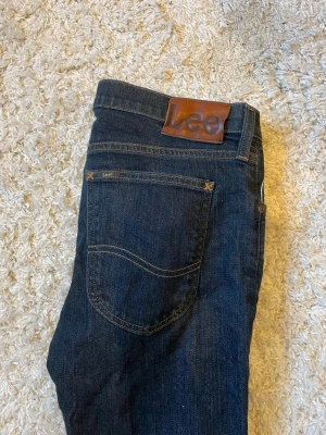 Mörkblå jeans från Lee - Snygga mörkblå jeans från Lee med klassisk femficksdesign och raka ben. Jeansen har en tydlig Lee-lapp i brunt läder bak i midjan och är tillverkade i slitstark denim. Perfekta för en avslappnad och trendig look.