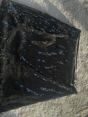 Svart paljettkjol från Amisu - Snygg svart kortkjol från Amisu täckt med glittriga paljetter som reflekterar blått och silver. Kjolen har en tight passform och är perfekt för dig som vill sticka ut. Materialet är syntet och kjolen har en slät insida för extra komfort.