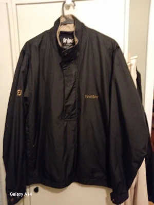 Svart vindjacka från FootJoy - Snygg svart vindjacka från FootJoy med diskreta loggor på bröstet och ärmen. Jackan har hög krage, dragkedja med vindslå och är tillverkad i ett lätt, vattenavvisande material. Perfekt för blåsiga dagar och sportiga outfits.