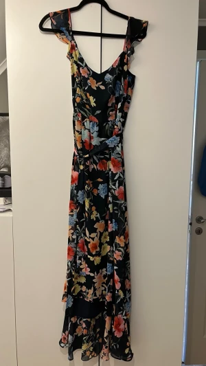 Blommig långklänning - Snygg långklänning med blommigt mönster i färger som blå, röd, gul och orange på mörk botten. Klänningen har volangdetaljer vid axlarna, v-ringad hals och markerad midja. Perfekt för sommarens alla tillfällen.