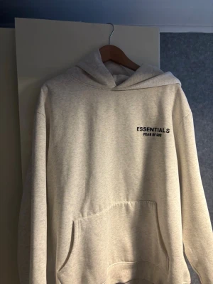 Essentials Fear of God hoodie grå - Snygg ljusgrå hoodie från Essentials Fear of God med svart logga tryckt på både bröstet och ryggen. Hoodien har en stor känguruficka framtill och en klassisk huva. Tillverkad i mjukt bomullsmaterial som är perfekt för en avslappnad streetwear-look.