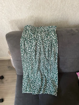 Grön blommig midikjol - Säljer en grön midikjol med vitt småblommigt mönster och resår i midjan. Kjolen har en avslappnad passform och är gjord i ett lätt och luftigt tyg, perfekt för vår och sommar. Superfin att matcha med en enkel topp eller hoodie.