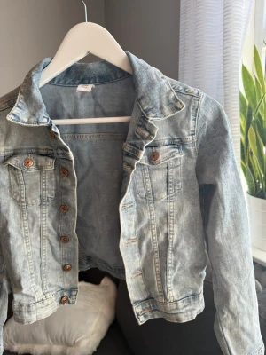 Ljusblå klassisk jeansjacka - Säljer en ljusblå jeansjacka med bronsfärgade knappar och två bröstfickor med lock. Jackan har en klassisk krage och raka ärmar. Perfekt att slänga över en hoodie eller t-shirt för en avslappnad look.