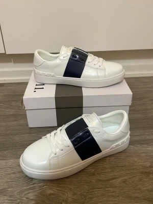 Valentino open skor - Snygga vita sneakers från Valentino Garavani med bred marinblå detalj över snörningen. Skorna har rund tå, platt sula och är tillverkade i skinn. Perfekta för dig som vill ha en clean och exklusiv look.