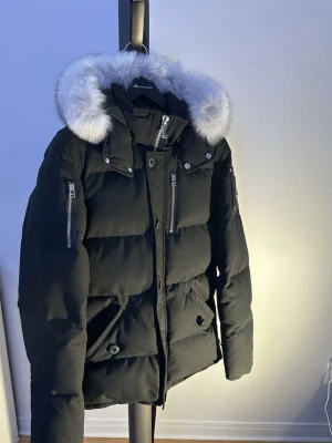 Svart dunjacka Moose Knuckles - Säljer en svart dunjacka från Moose Knuckles med stor fluffig vit päls. Jackan har flera dragkedjeförsedda fickor, tryckknappar och en snygg logga på ärmen. Perfekt för kalla vinterdagar och när det kallt om nätterna och riktigt stilren design. OBS påse från Moose kommer med.