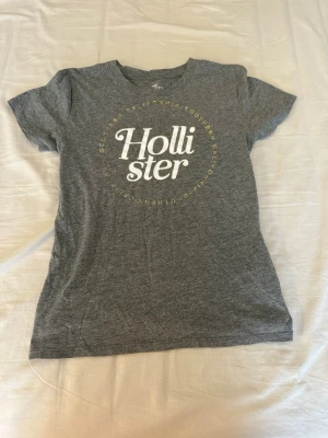 Grå Hollister t-shirt med tryck - Snygg grå t-shirt från Hollister med vitt tryck på bröstet och rund hals. T-shirten har korta ärmar och är tillverkad i mjuk bomullsmix som känns skön mot huden. Perfekt för en avslappnad och stilren look.